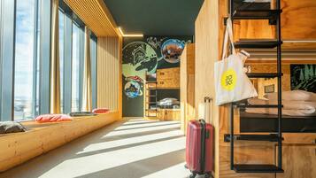 Ein modernes Hotelzimmer mit großen Fenstern, einem Stockbett, einem Koffer und einer Holzwand mit Stadtbild-Mural.