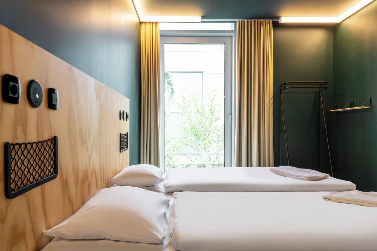 Une chambre d’hôtel avec deux lits, une tête de lit en bois, une fenêtre avec des rideaux et un porte-serviettes.