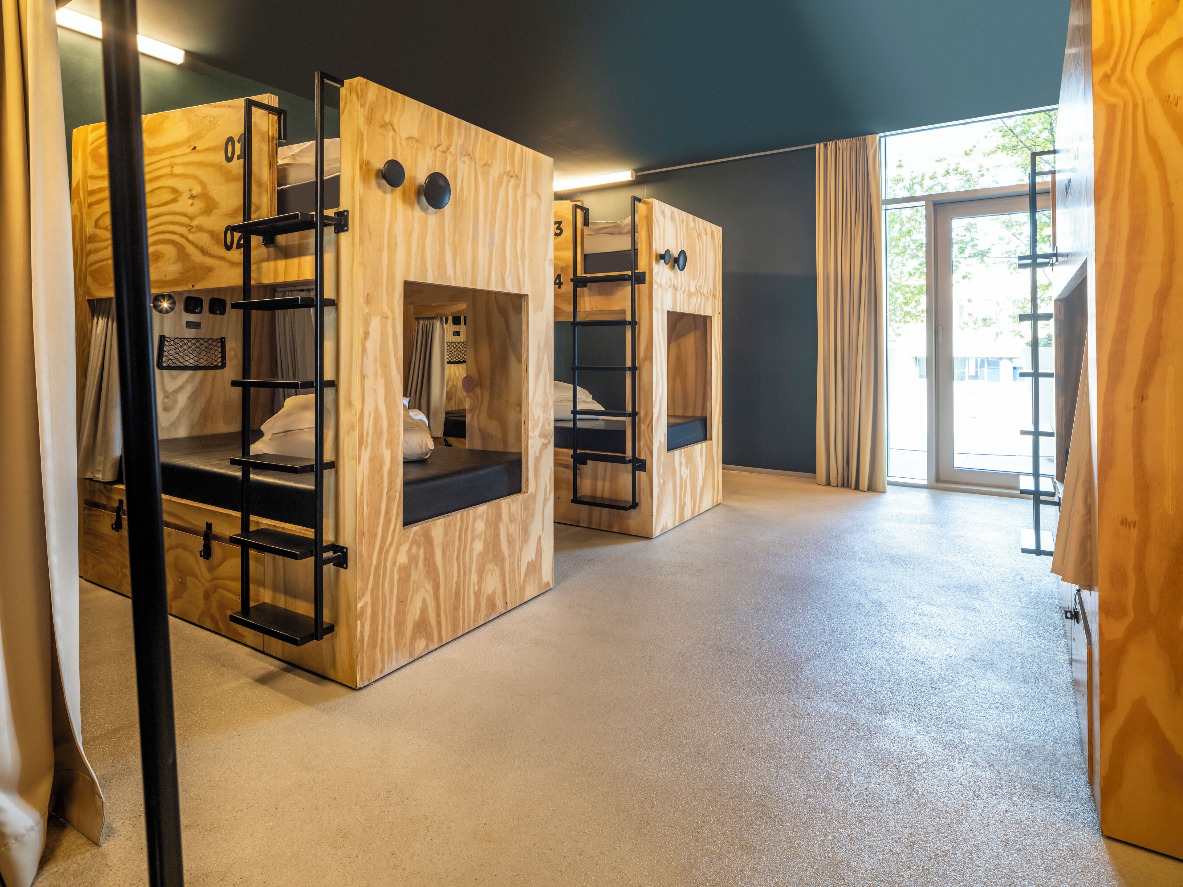 A modern, minimalist hotel room with bunk beds, a small desk, and a large window offering a view of the outdoors.