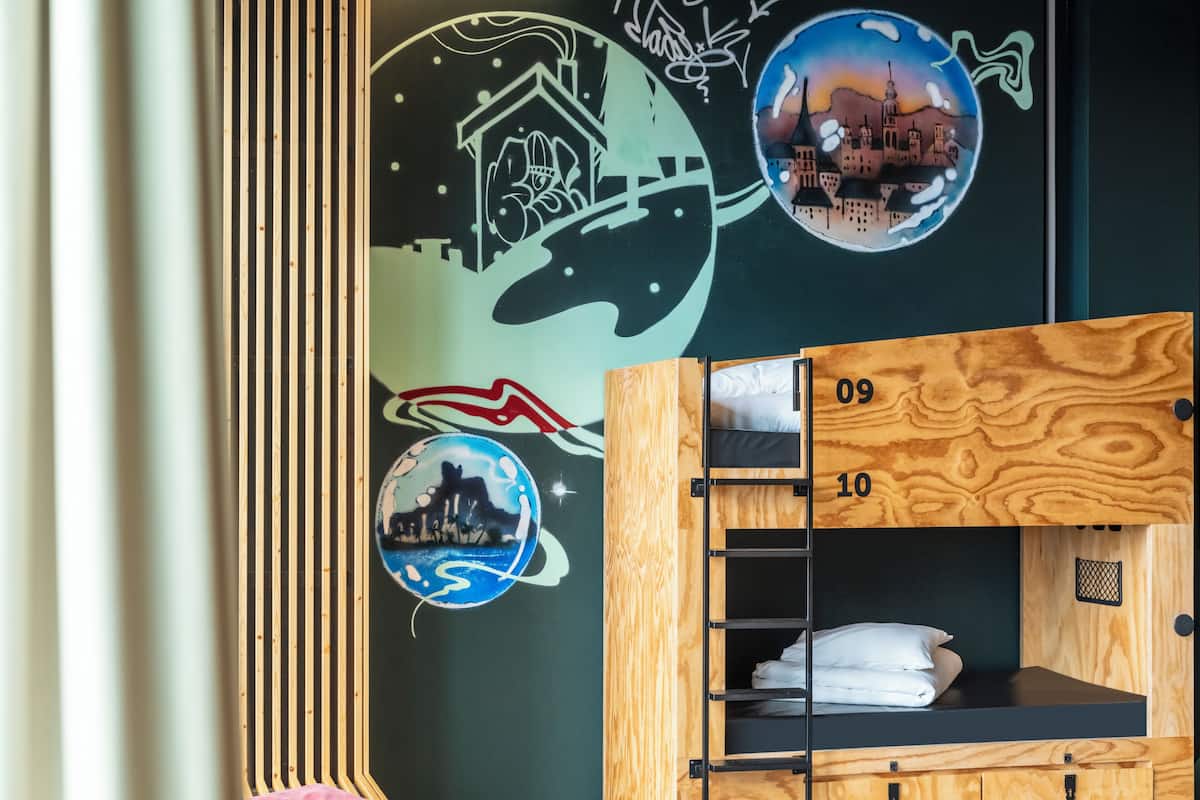 Un lit superposé avec une structure extérieure en bois et un matelas noir, un oreiller rose et un mur décoré de graffitis.