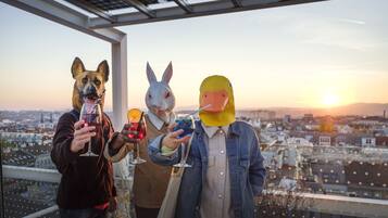 Three people in animal masks holding drinks on a balcony with a city view.