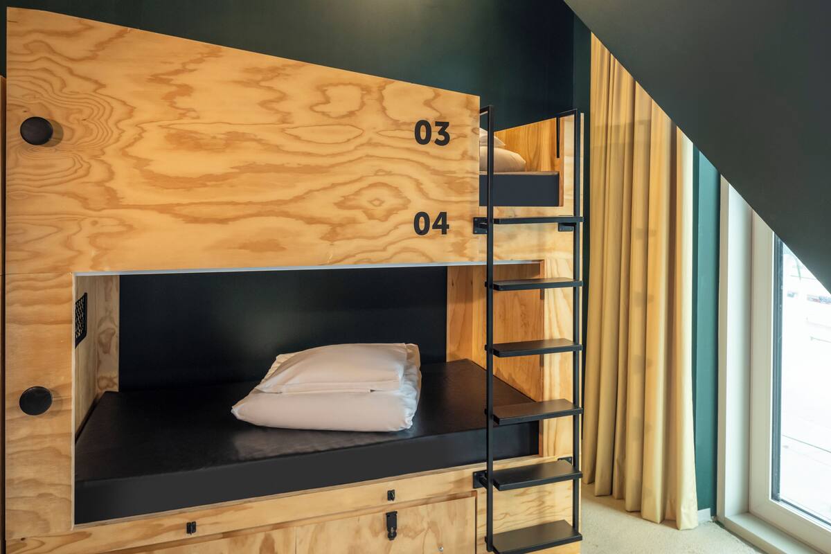 Un lit superposé avec une structure extérieure en bois et des étagères noires, portant les numéros 03 et 04, dans une pièce dotée d’une fenêtre et de rideaux.