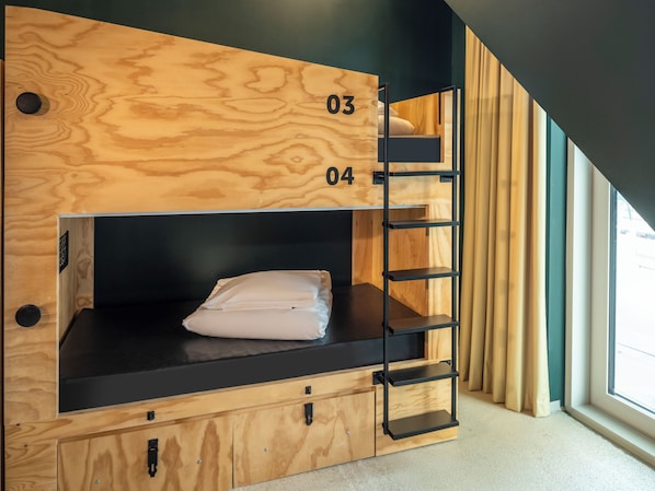Un lit superposé avec une structure extérieure en bois et des étagères noires, portant les numéros 03 et 04, dans une pièce dotée d’une fenêtre et de rideaux.