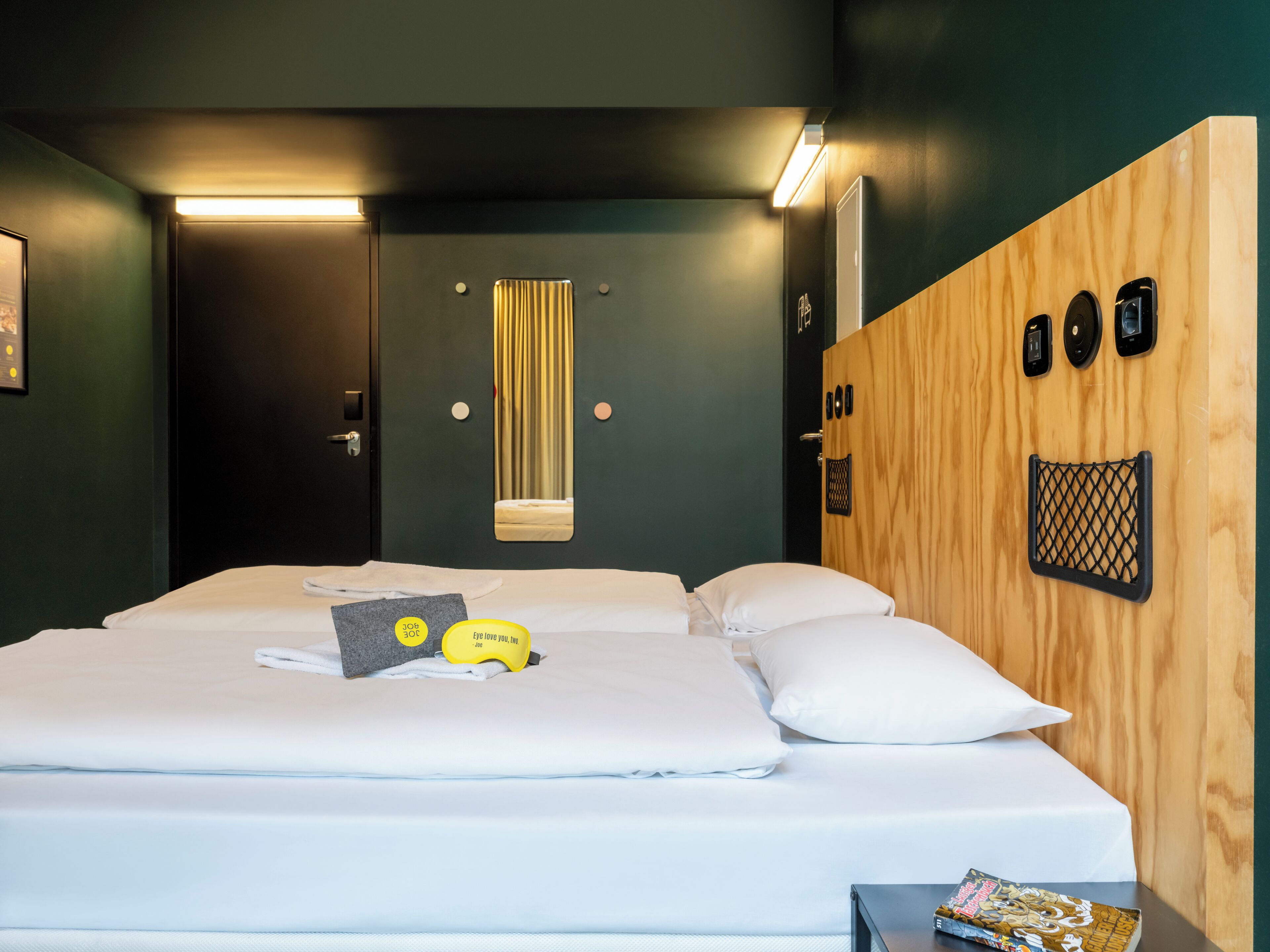 Quarto de hotel com cama, cabeceira de madeira, espelho e uma mesinha com uma revista.