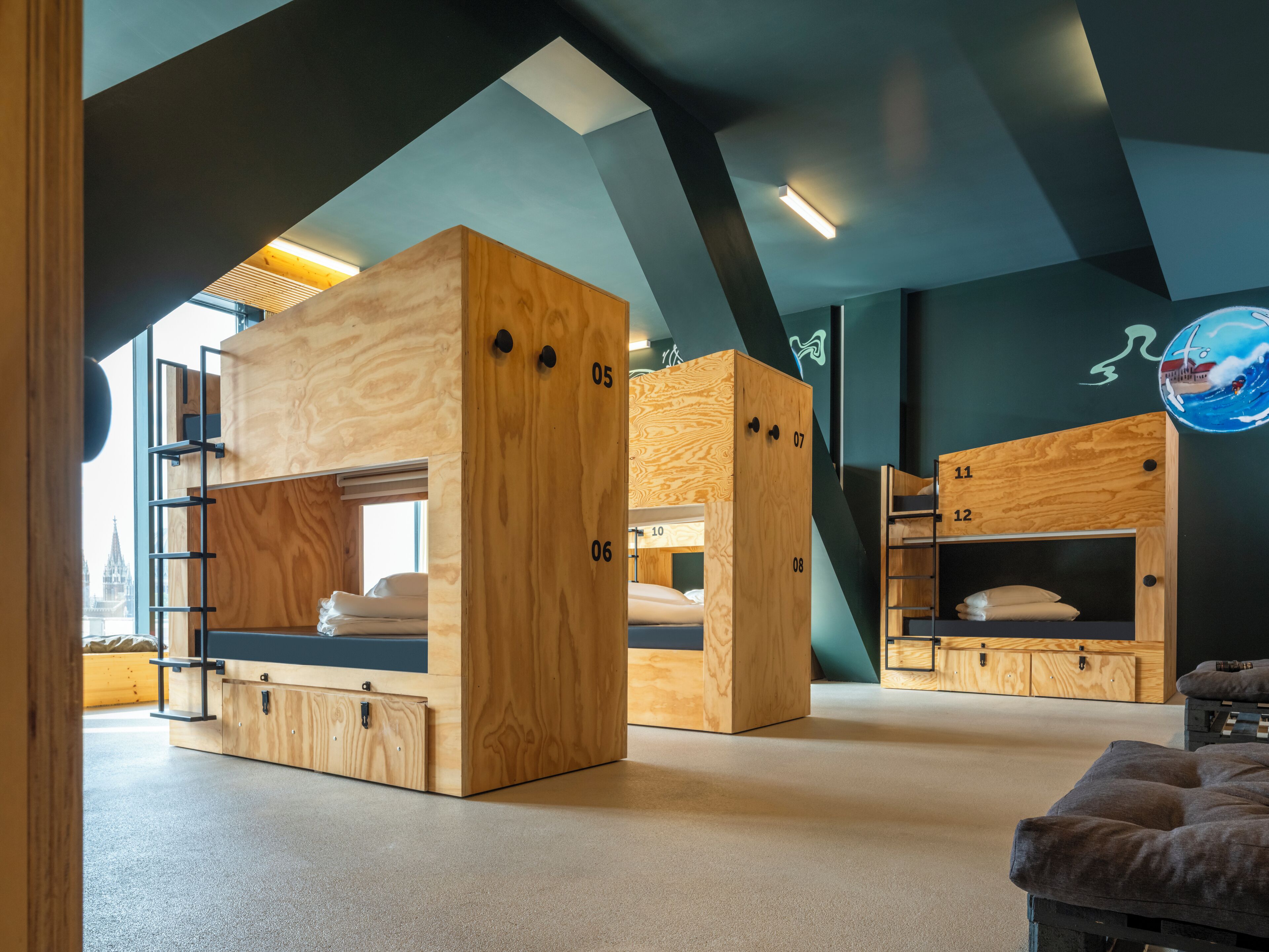 Een moderne, meerlaagse slaapzaal met houten stapelbedden, genummerde compartimenten en uitzicht op een stadslandschap door een groot raam.