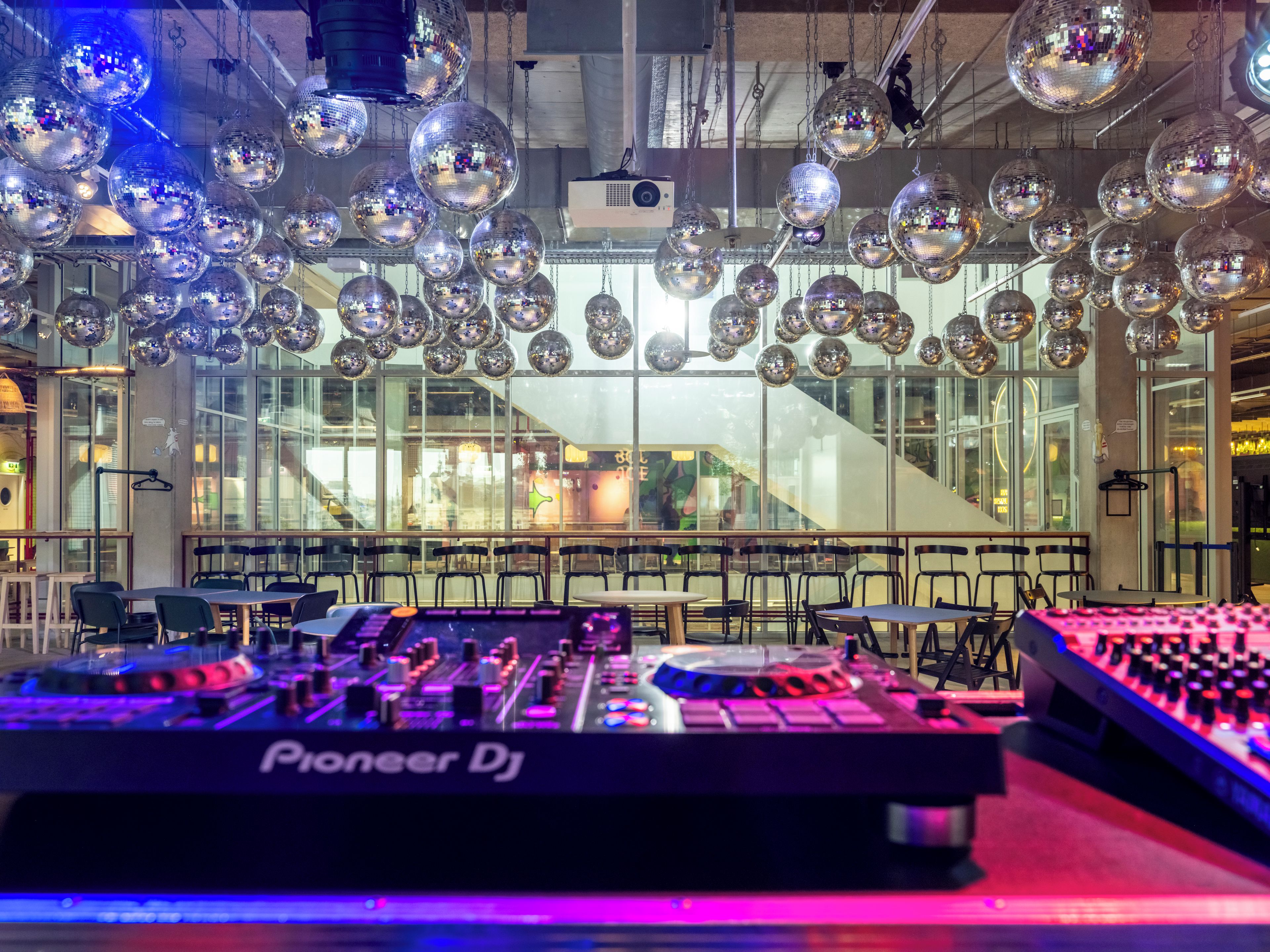 A DJ booth with Pioneer DJ equipment, surrounded by tables and chairs, and disco balls hanging from the ceiling.