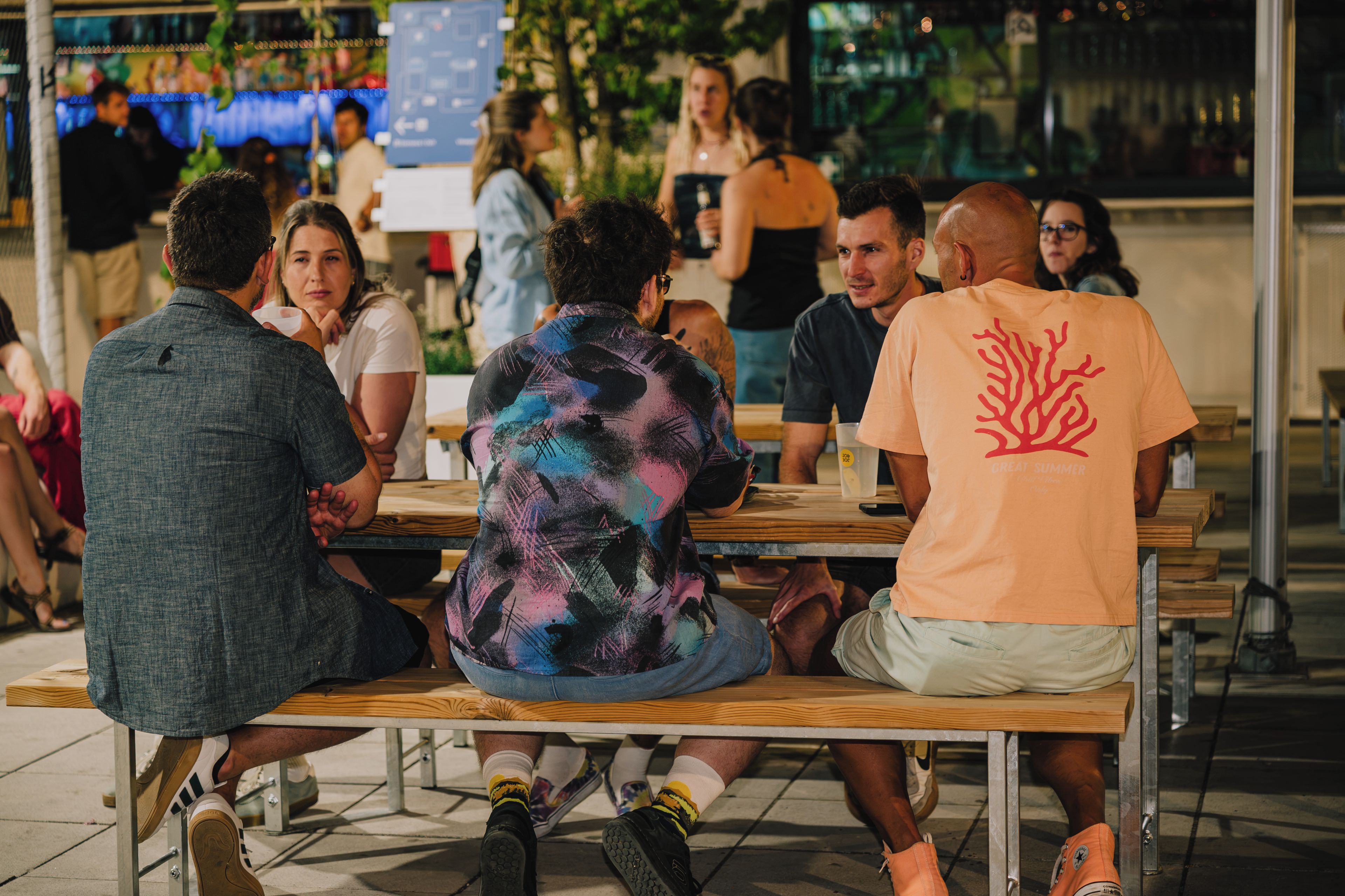 Un groupe de personnes est assis à des tables en bois à l’extérieur, probablement dans un café ou un restaurant.