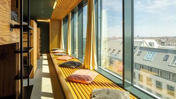 A row of wooden benches with cushions, a wooden bookshelf, and large windows offering a city view.