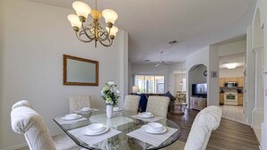 A dining area with a glass table, white patterned chairs, a chandelier, a mirror, and a view into the living room.