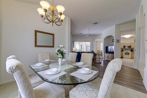 A dining area with a glass table, white patterned chairs, a chandelier, a mirror, and a view into the living room.
