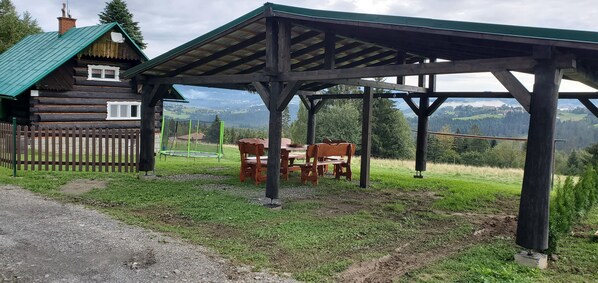 Uma cabana de madeira rústica com telhado verde, um deque de madeira e uma área coberta com bancos e uma mesa.