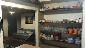 Ein gemütliches Wohnzimmer mit einem grünen Sofa, einem Esstisch und einer Küchenzeile mit Regalen voller Geschirr und Gläsern.