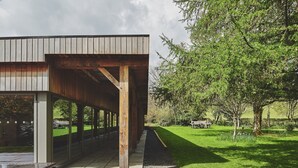 A modern building with a covered outdoor area, a stone pathway, and a grassy landscape with trees. - Honister Cottage - Luxury Cottages (Kendal)
