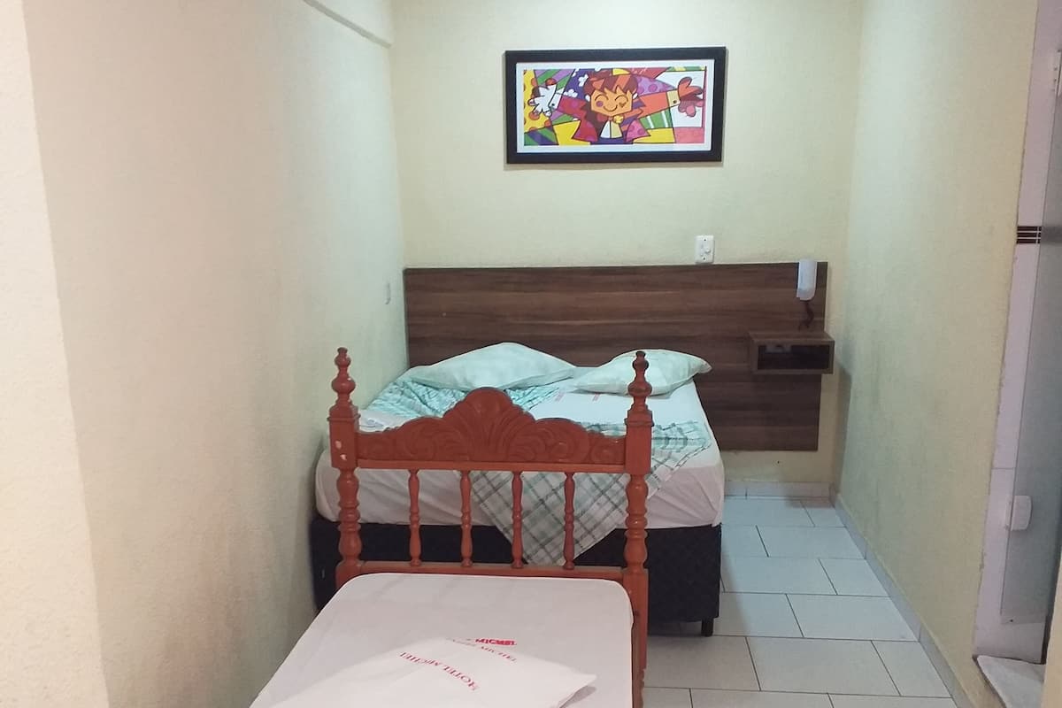 A small, single-bed room with a wooden headboard, a ceiling fan, and a colorful painting on the wall.