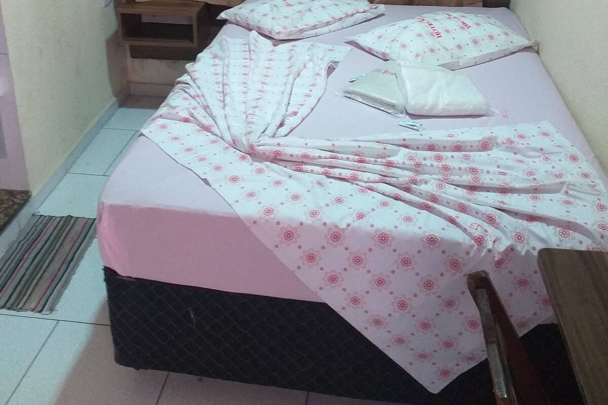 A small, single-bed room with a pink bedspread, white pillows, and a wooden headboard. There is a window with beige curtains, a wall-mounted air conditioner, and a small wooden shelf.