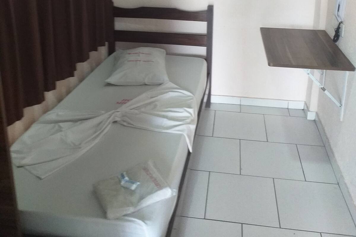 A small, single-bed room with a wooden headboard, a small table, and a window with a picture frame.