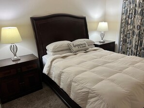 Een net opgemaakt bed met een sierkussen waarop staat 'geluk is zelfgemaakt', omringd door twee bedlampen en een nachtkastje van donker hout.