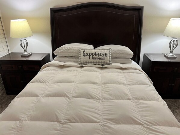 Een net opgemaakt bed met een gewatteerde deken en twee kussens, waarvan één met de tekst 'happiness is homemade'. Aan beide zijden van het bed staan nachtkastjes met lampen.