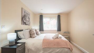 A bedroom with a bed, pillows, a nightstand with a lamp, a window with curtains, and a painting on the wall.