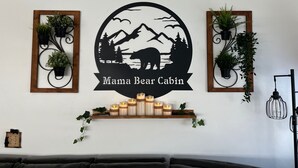 A wall with a circular logo reading "Mama Bear Cabin," flanked by two plant holders with greenery, and a shelf with lit candles. - MAMA BEAR CABIN -RUSTIC LOG  Cabin Retreat -  Boat Parking - All Size Groups (Garden City)