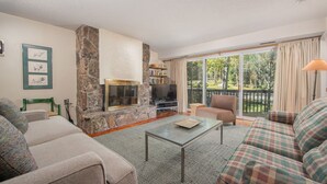 A living room with a stone fireplace, a sofa, a coffee table, and a view of a garden through large sliding glass doors. - JHRL - Slope-side Whiteridge B1 Condo - Walk to Tram (Teton Village)