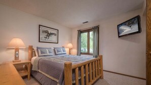 A bedroom with a wooden bed, two bedside tables with lamps, a window with curtains, and a wall-mounted TV.