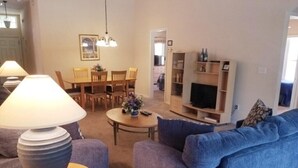 Un salon avec un canapé bleu, une table basse ronde et une lampe. On y trouve également un coin repas avec une table et des chaises, ainsi qu’un téléviseur posé sur un meuble.