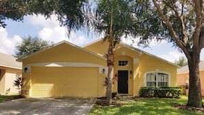 Une maison jaune de plain-pied, avec un garage, une porte d’entrée et un petit jardin devant, agrémenté d’un arbre et de quelques arbustes.