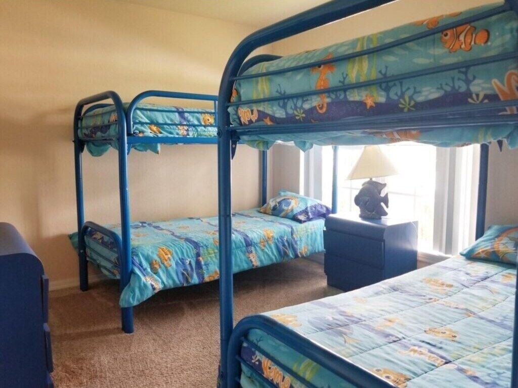Ein Zimmer mit drei Etagenbetten, jeweils mit blauem Gestell und farbenfroher Bettdecke. In der Ecke steht ein kleiner Tisch mit einer Lampe und einem Stuhl.