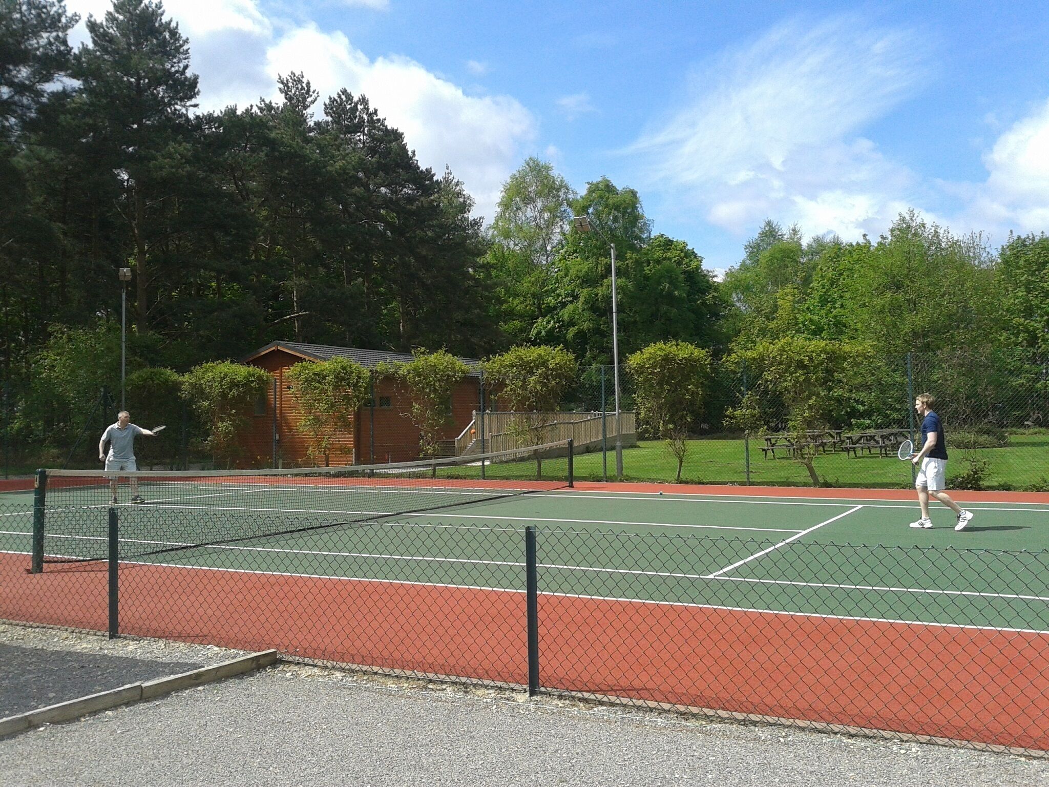 Una cancha de tenis al aire libre con red, rodeada de árboles y un pequeño edificio.