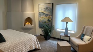 Ett hotellrum med en säng, en stol, en lampa, en tavla och en öppen spis.