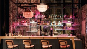 Una zona de bar con varias estanterías llenas de botellas y vasos, tres taburetes y una lámpara de araña grande.