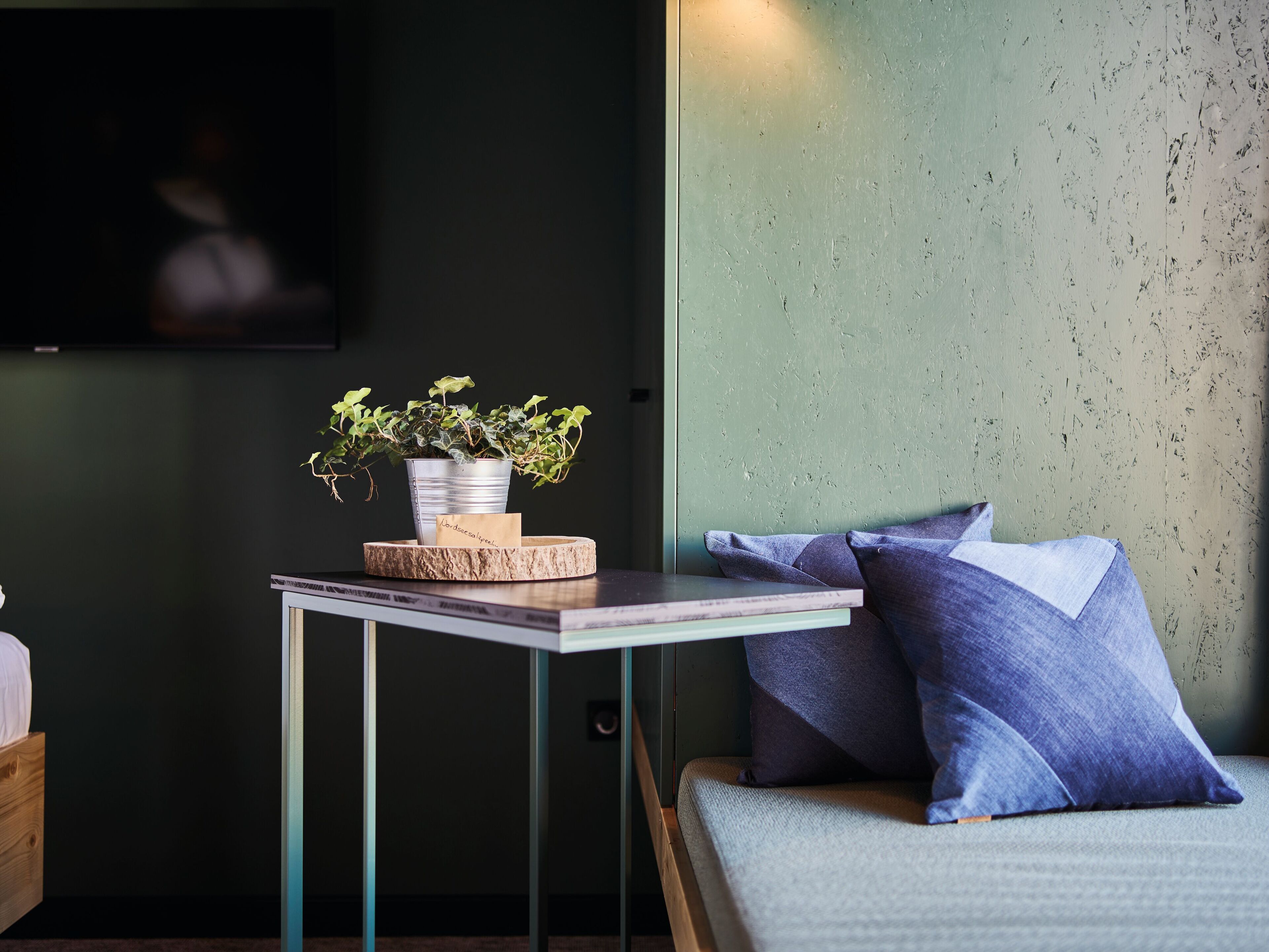 Ein kleiner Tisch mit Pflanze, Notizbuch und Stift. Ein Bett mit Kissen und ein an der Wand befestigter Fernseher.