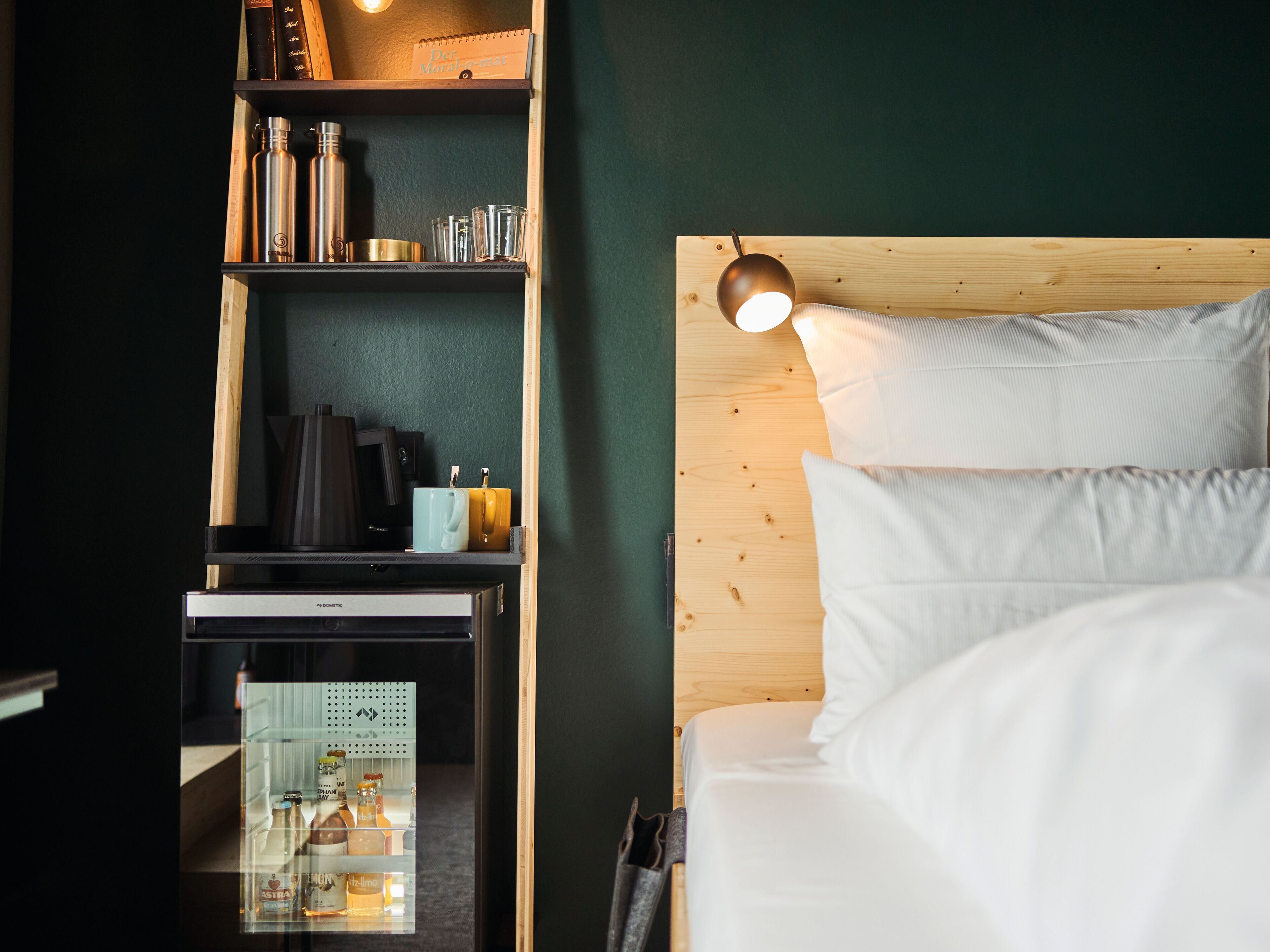 Une chambre d’hôtel avec une tête de lit en bois, un lit recouvert de draps blancs, une étagère avec des livres et des bouteilles, et un petit bureau avec un ordinateur et une lampe.