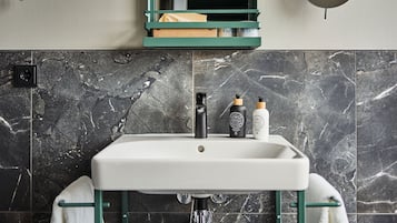 A bathroom with a white sink, dark marble tiles, a green-framed mirror, and two toiletry bottles on the sink.