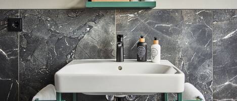 A bathroom with a white sink, dark marble tiles, a green-framed mirror, and two toiletry bottles on the sink.