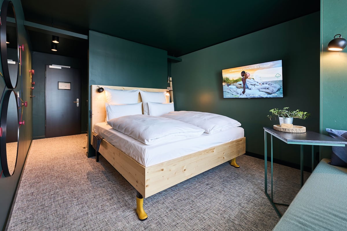 Un dormitorio moderno con una cama de madera, una mesita de noche pequeña con una planta y un televisor montado en la pared mostrando un mensaje de bienvenida.