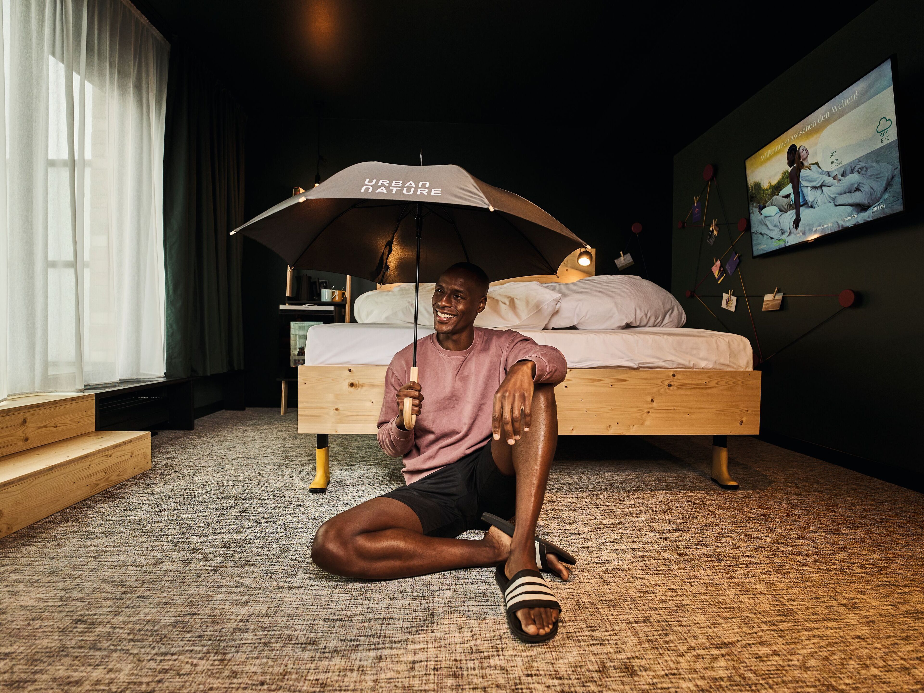 A bedroom with a bed, a person sitting on the floor holding an umbrella, and a TV on the wall displaying a weather forecast.