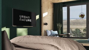 Un dormitorio con cama, un televisor empotrado mostrando “URBAN NATURE”, una ventana con vistas al campo y una mesita de noche con un jarrón de flores.