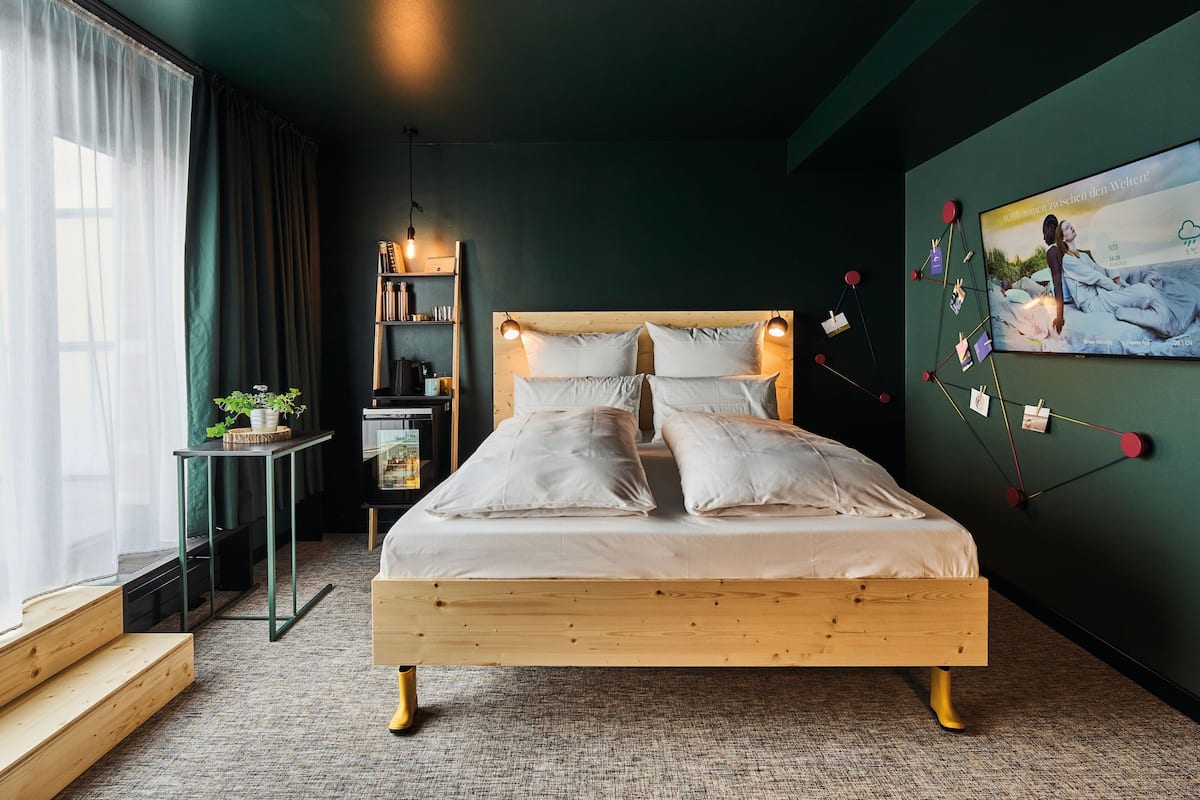 Un dormitorio moderno con una cama de madera, una mesita de noche pequeña, una estantería con libros y una planta, y un televisor empotrado que muestra una imagen panorámica.