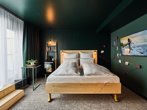 Un dormitorio moderno con una cama de madera, una mesita de noche pequeña, una estantería con libros y una planta, y un televisor empotrado que muestra una imagen panorámica.