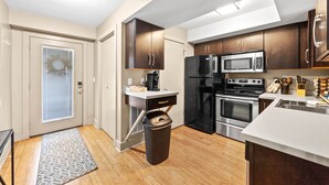 A compact kitchen with dark wooden cabinets, a stainless steel refrigerator, a microwave, and an oven. There is a small countertop with a coffee maker and a trash can. - The Lofts of Downtown (540 N 2nd St, #105) (Clarksville)