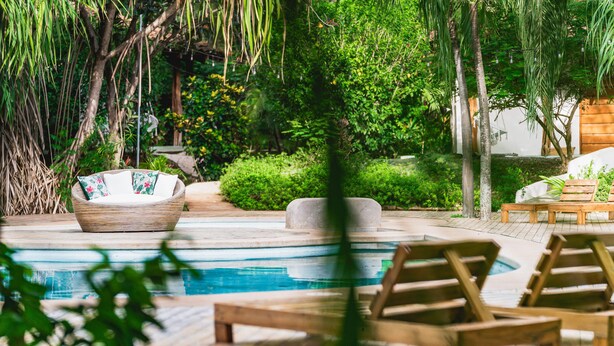 Area piscina con lettini in legno, una sedia in vimini con cuscini e verde lussureggiante.