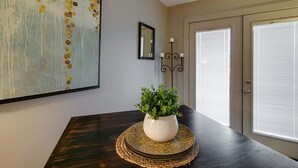 Un comedor con una mesa de madera oscura, un florero blanco con plantas verdes, un cuadro abstracto grande, un espejo y una puerta con persianas.