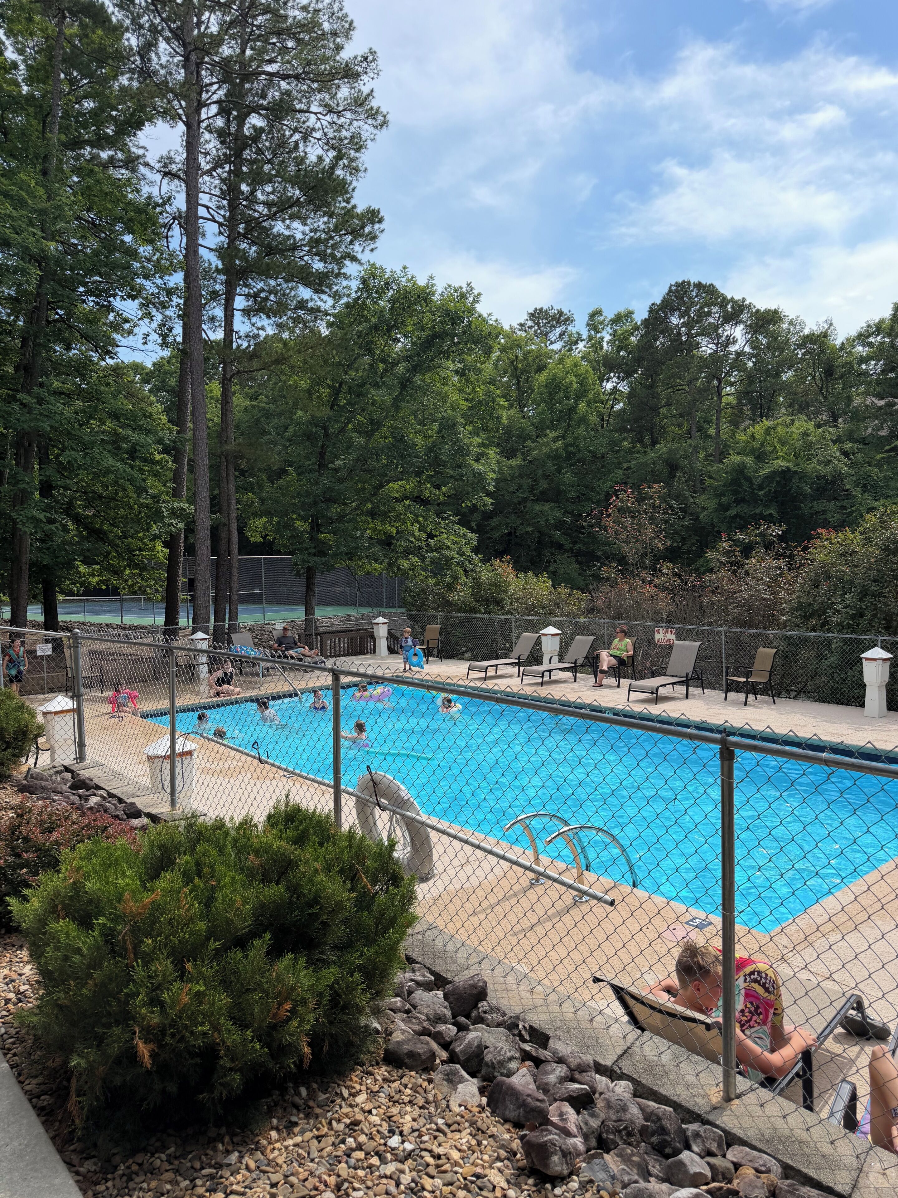 A swimming pool surrounded by a fence, with trees in the background and a few people around.