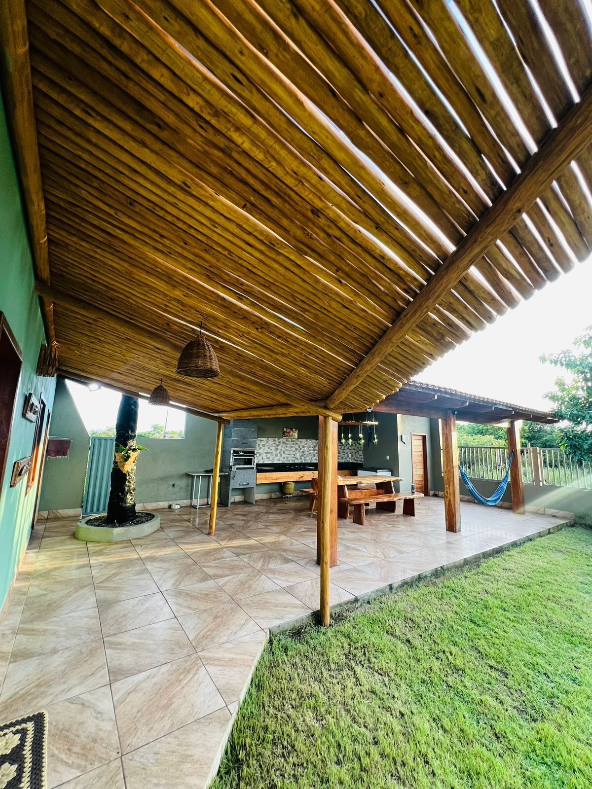 A covered outdoor patio with a tiled floor, a wooden pergola, a hammock, and a kitchen area with a stone backsplash.