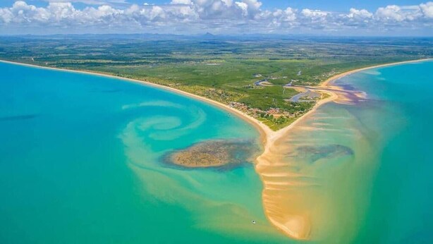 Vista aérea de una zona costera con aguas turquesas cristalinas, playas de arena y un pequeño asentamiento con calles y edificios visibles.