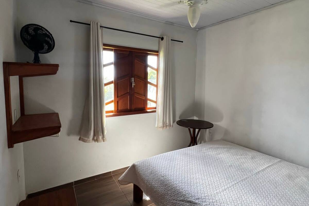 Un dormitorio con piso de madera, una cama con cubrecama blanco texturizado, un ventilador de techo, una mesita y una ventana con cortinas.