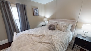 A bedroom with a bed, a nightstand, a lamp, and a window with curtains. - Beautiful Bungalow nestled in Nelsonville near Hocking Hills (Nelsonville)