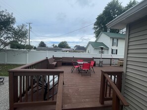 A wooden deck with outdoor furniture, a metal trash can, and a white house in the background. - Beautiful Bungalow nestled in Nelsonville near Hocking Hills (Nelsonville)